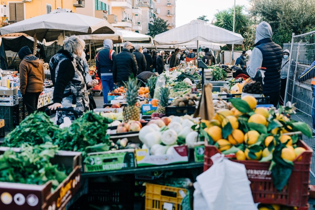 People walk through an outdoor market in cold weather wearing jackets and vests, browsing stacks of produce.