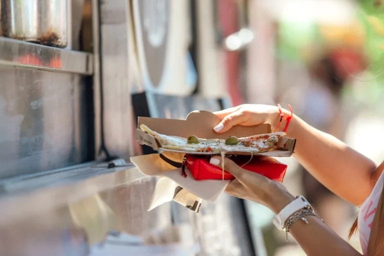 A close-up shot of a person holding a slice of pizza from a food truck.