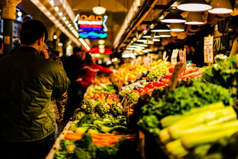 A person walks through a covered farmers market next to stacks of produce with neon signs overhead.