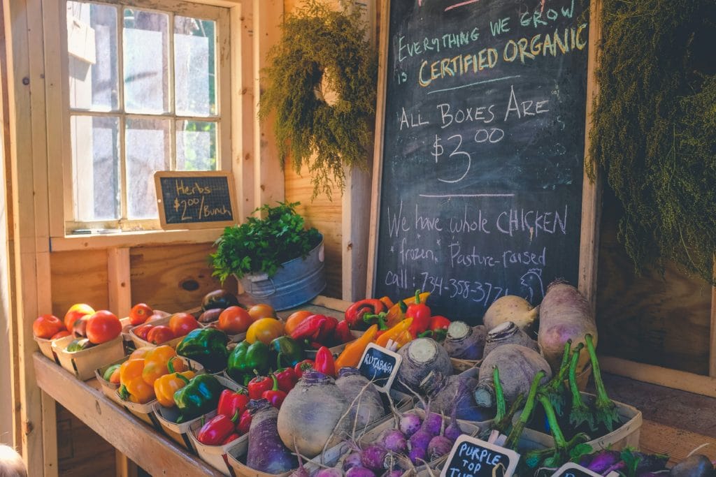 A small display of fruits and vegetables at an indoor market with chalkboard signage displaying prices and product names.