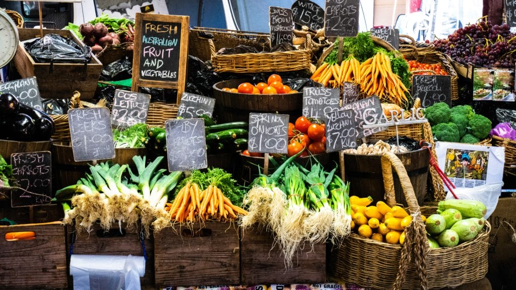 Various vegetables displayed in wooden crates and baskets at an outdoor market with chalkboard signage.