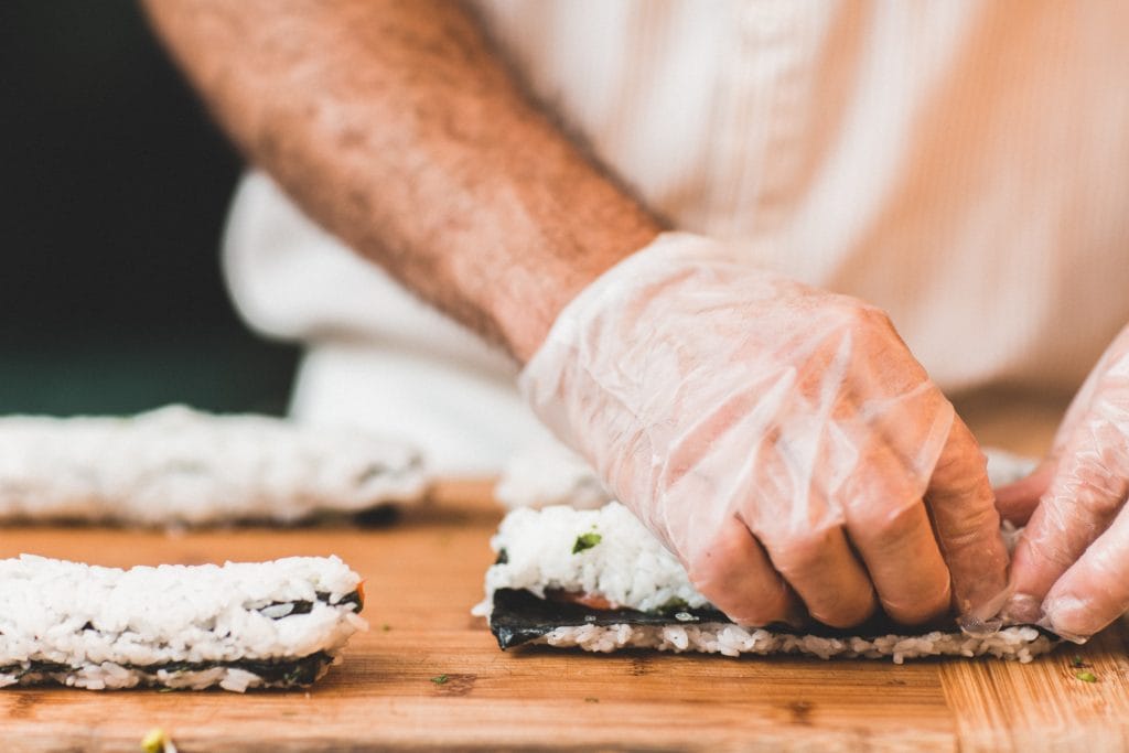 A close-up photo of a food service worker making sushi rolls with gloved hands.