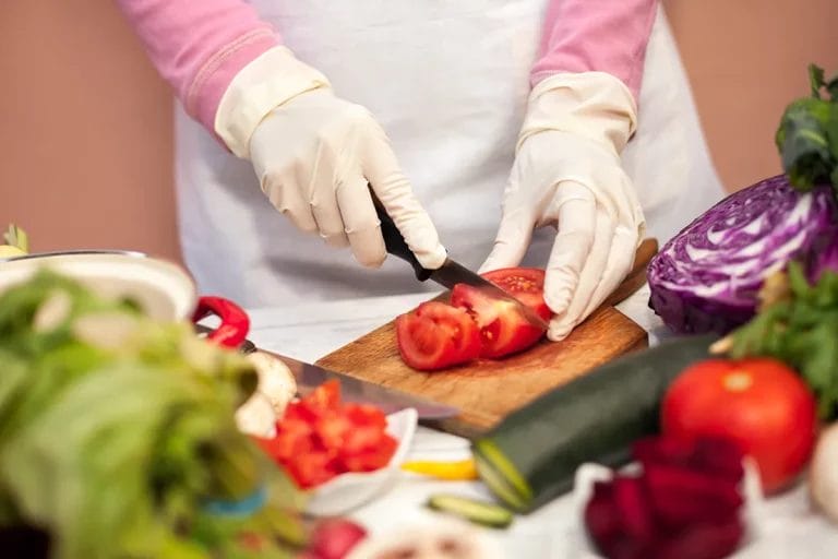 A person wearing food-safe disposable gloves slicing a tomato on a cutting board.