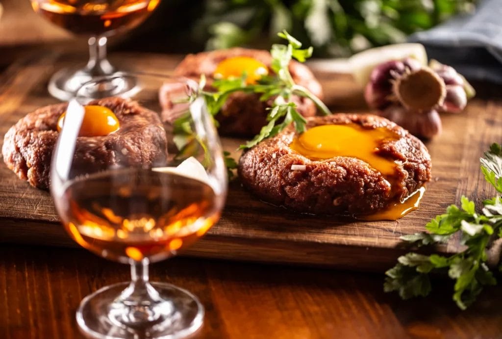 A glass of whiskey in front of a cutting board with beef tartare.