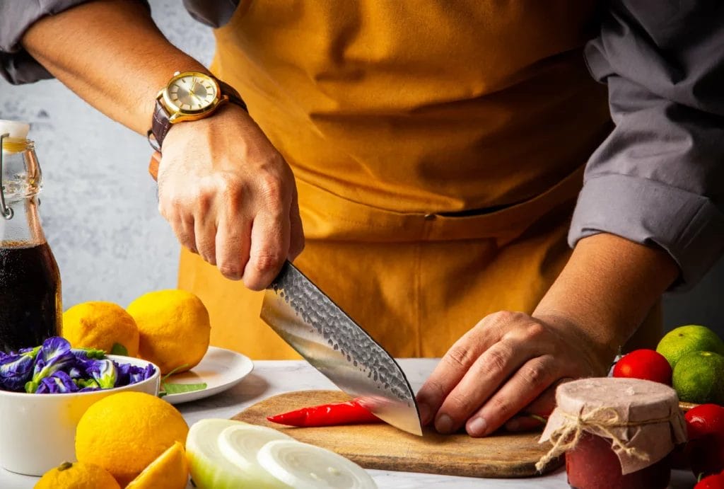 A chef in a mustard yellow apron chopping a red pepper on a cutting board surrounded by other ingredients.
