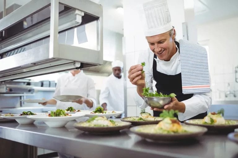 A smiling chef garnishes dishes in a kitchen.