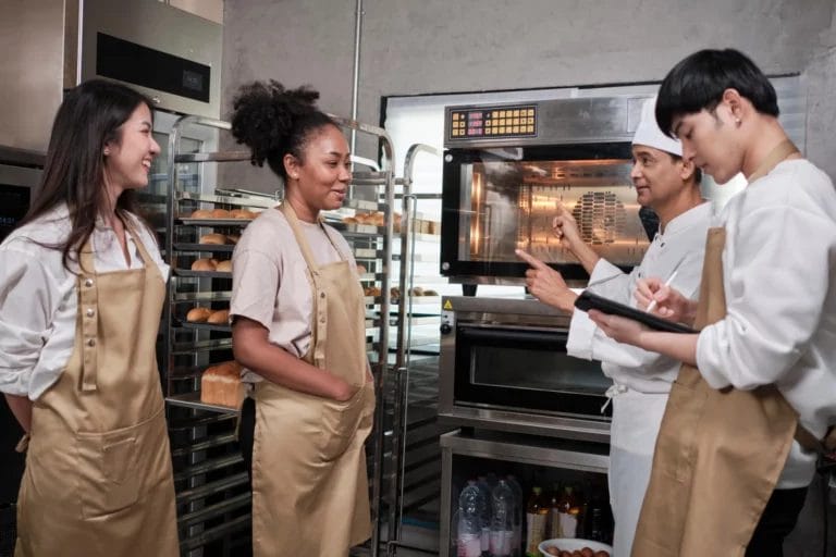 A baker teaches three students in tan aprons in a commercial kitchen next to bread racks and ovens.