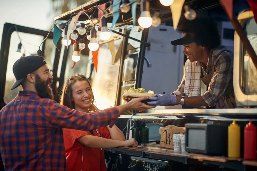 A woman working in a food truck hands a basket of food to a man and woman.
