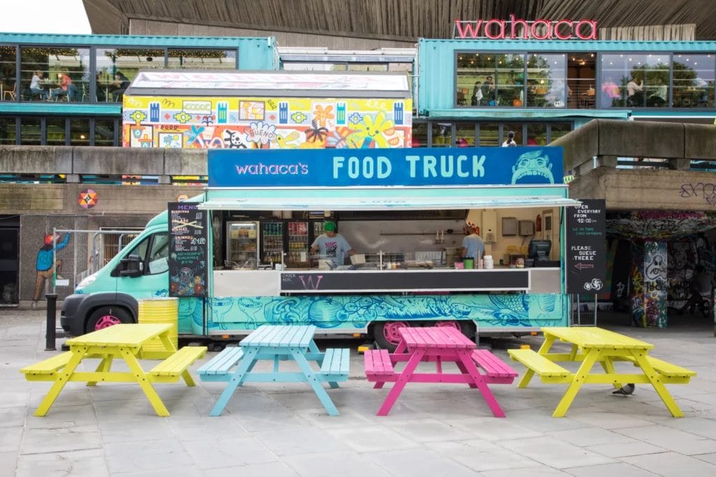 A turquoise food truck with the words "Wahaca's Food Truck" is open for business behind brightly colored yellow, blue, and pink picnic tables in a paved courtyard.