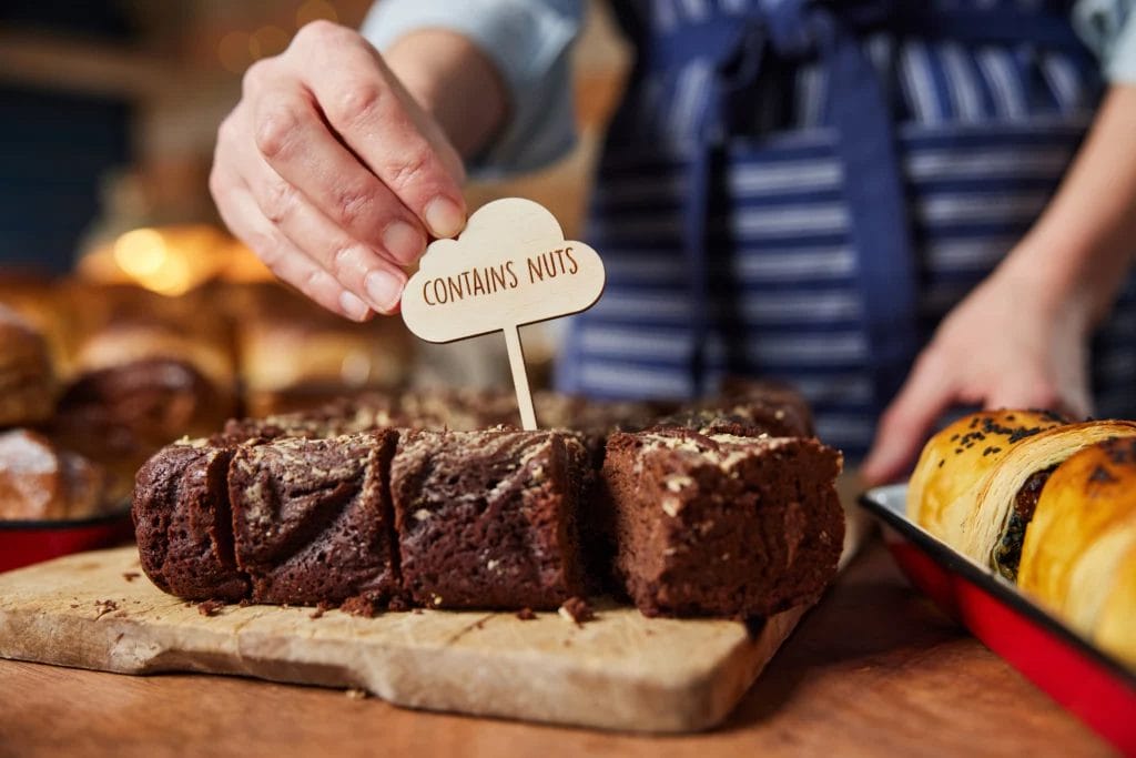 A baker in a blue striped apron places a small wooden sign that reads "Contains Nuts" in a display of chocolate baked goods.