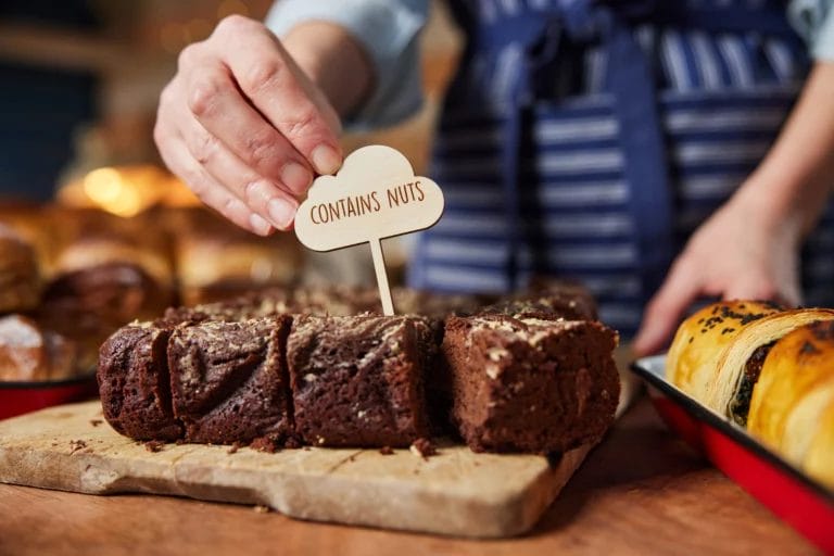 A baker in a blue striped apron places a small wooden sign that reads "Contains Nuts" in a display of chocolate baked goods.