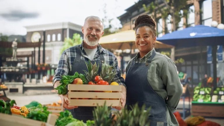 A man and woman stand in front of their produce booth at a farmers market wearing black aprons and smiling at the camera.
