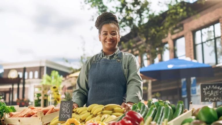 A woman wearing a green shirt and black apron stands behind her produce booth at a farmers market and smiles at the camera.