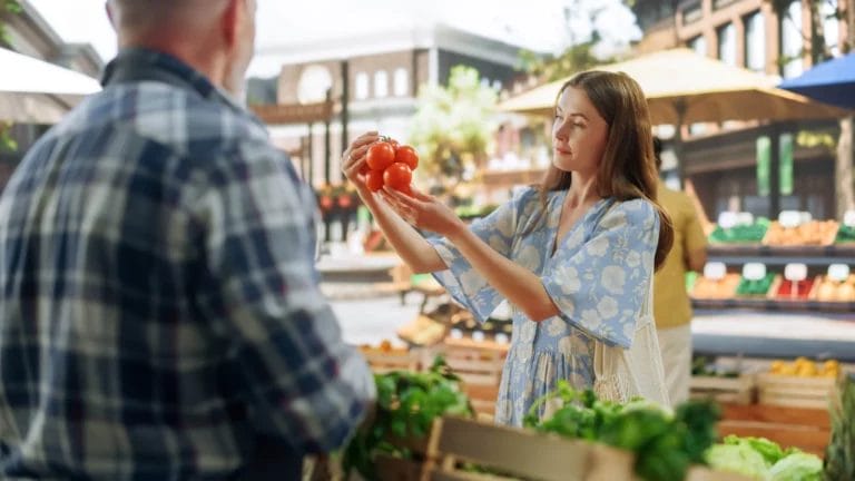 A young woman with long brown hair and a loose-fitting blue floral top inspects some tomatoes on the vine in front of a male farmers market vendor at his produce stall.