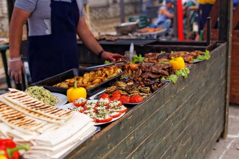 A food festival vendor booth displaying various foods including grilled flatbreads, meats, and stuffed bell peppers.