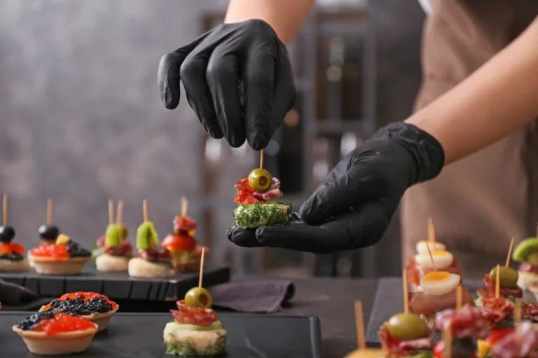 A caterer arranges a tiered charcuterie board with cured meats and artisan cheeses while wearing black sterile gloves.