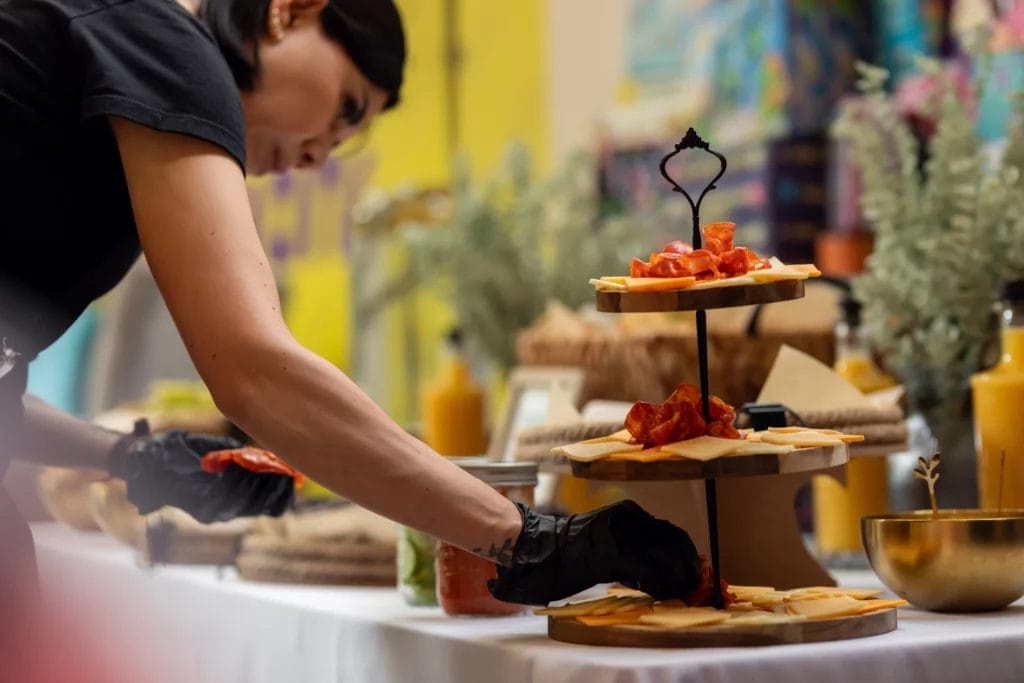 A caterer arranges a tiered charcuterie board with cured meats and artisan cheeses while wearing black sterile gloves.