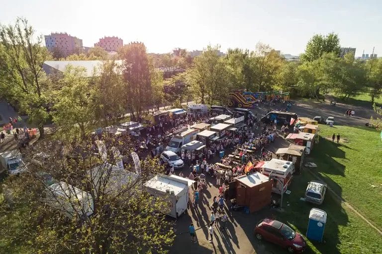 A food truck rally photographed from above on a sunny day. Crowds of people walk between the trucks ordering food.