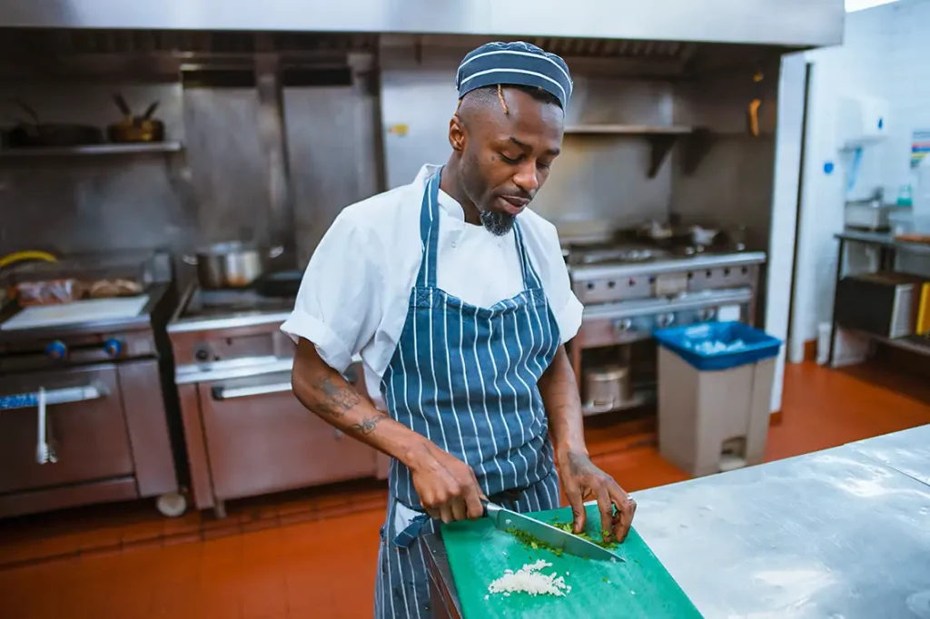 A chef wearing a blue and white striped apron dices vegetables on a cutting board in a commercial kitchen.