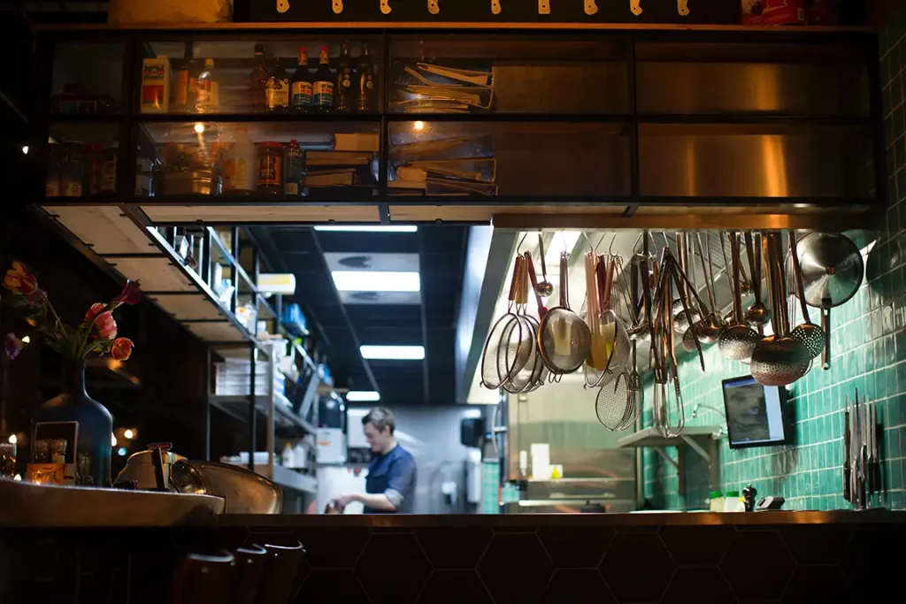 Strainers and ladles hang from overhead storage cabinets stocked with sauces and other ingredients in a commercial kitchen. In the background, a chef prepares food.