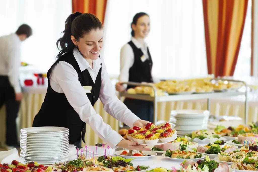 A catering employee smiles as she places a plate of pastries on a table full of food.