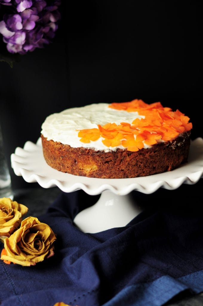 A single-layer cake on a white porcelain cake stand with orange flowers on top of the frosting.