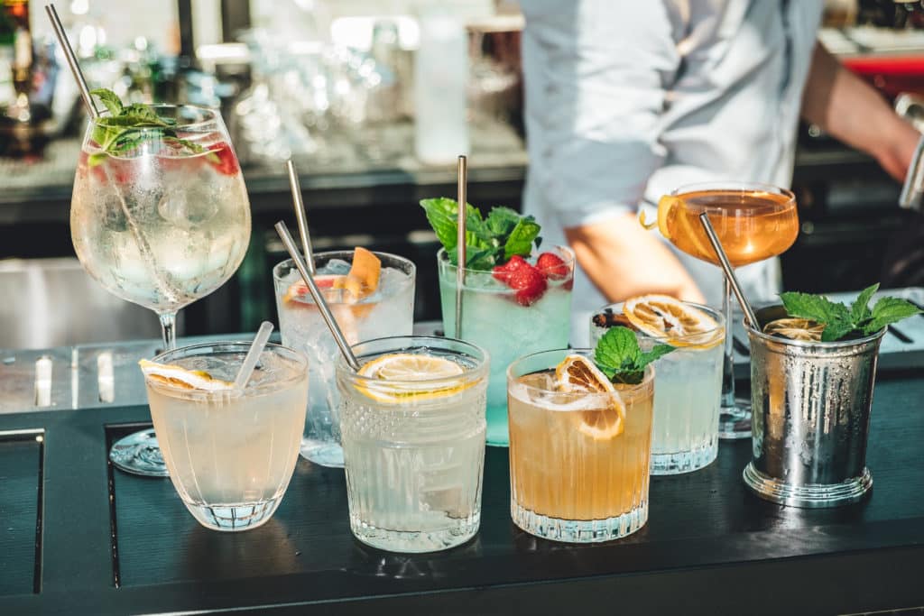 Nine different cocktails on a bar, each with different garnishes including citrus slices, berries, and mint leaves.