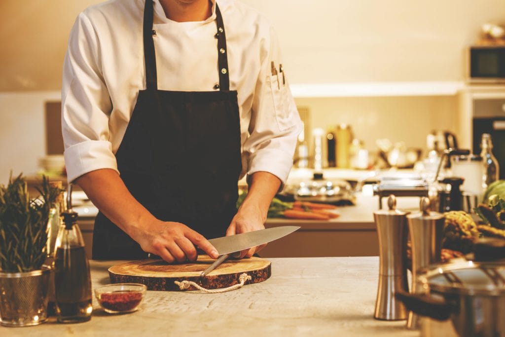 A personal chef wearing a black apron sharpening a knife in a home kitchen.