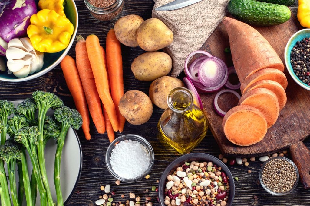 An overhead view of resh vegetables, salt, seeds, and oils on a rustic wooden countertop.