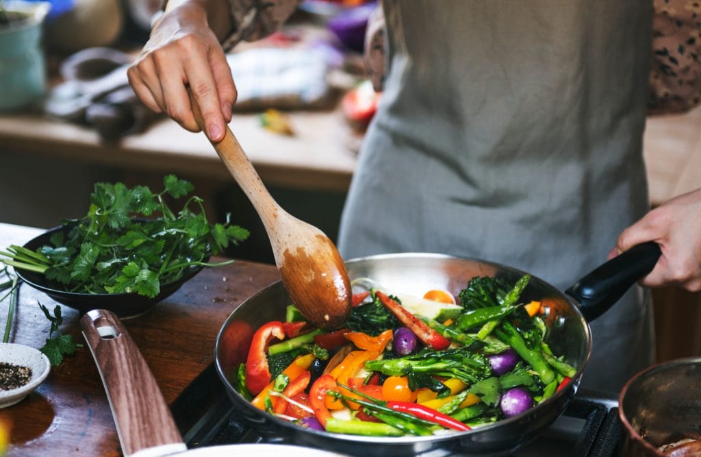 A close-up shot of a chef sprinkling ground red chili pepper into a frying pan filled with vegetables.