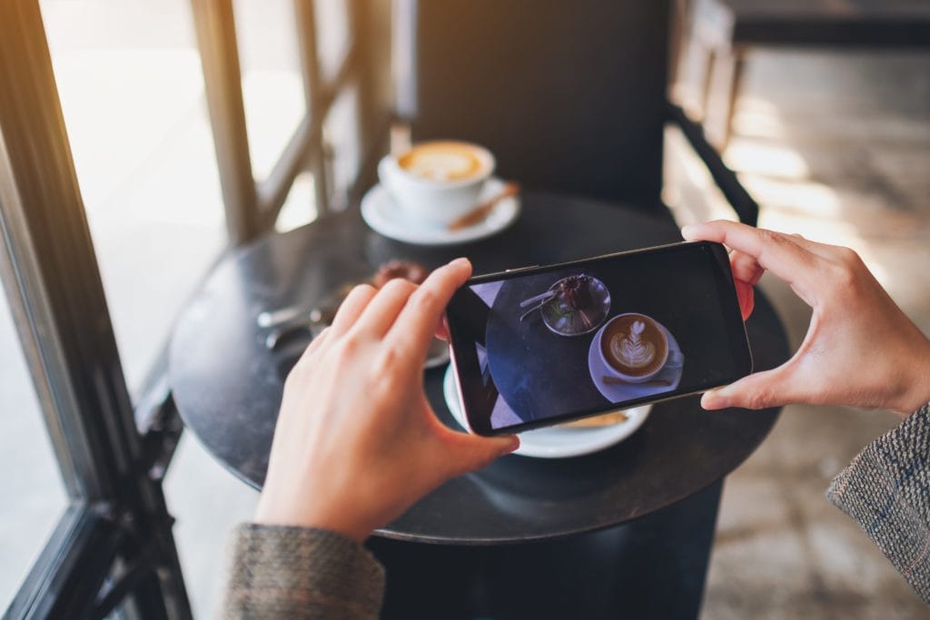 A person taking a picture of a latte and a snack with their smartphone.