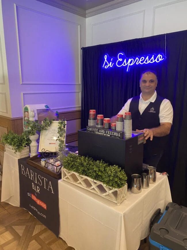 A man stands behind the Si, Espresso booth next to a coffee machine with a neon sign behind him.