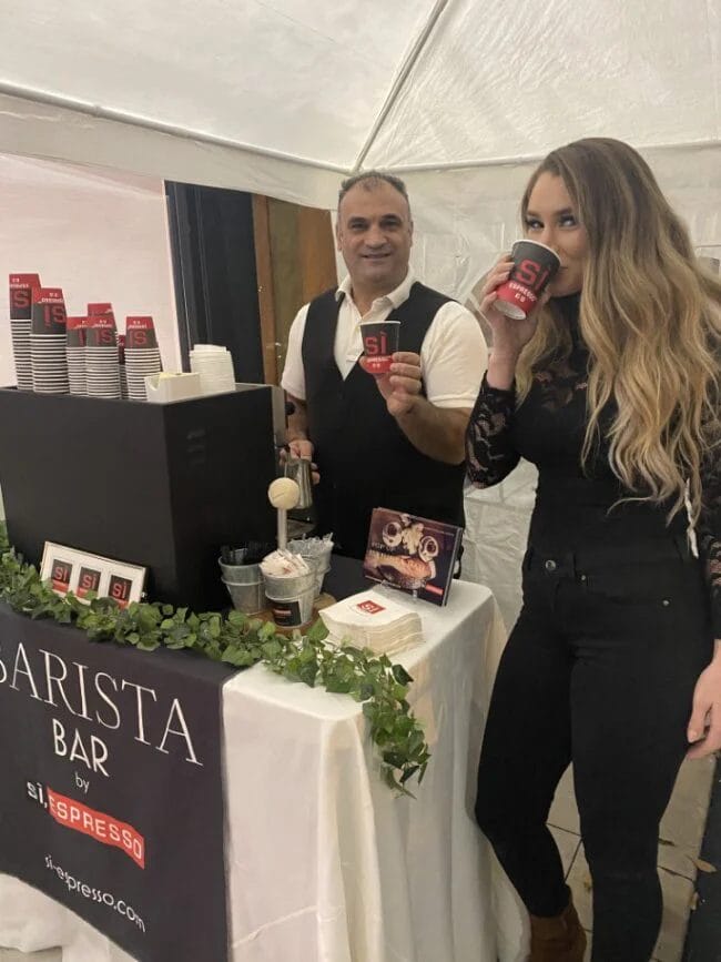 A man stands behind the Si, Espresso booth while a woman sips on a cup of coffee.