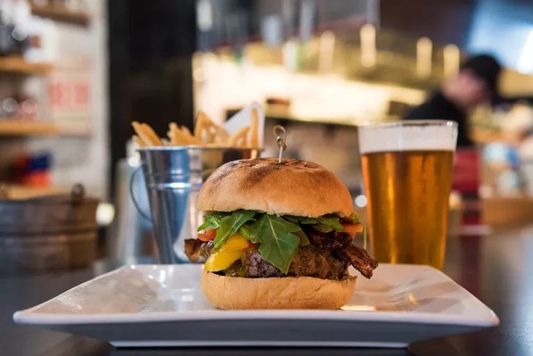 A burger and a metal bucket of fries with a glass of beer on the side.