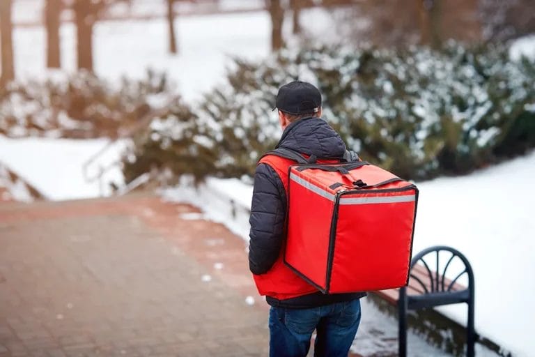 A food delivery person walking on a snowy sidewalk with an insulated red bag.