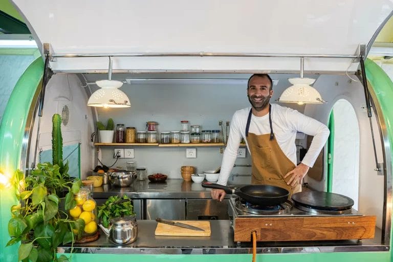 A man in a brown apron smiles at the camera from inside a green food truck with shelves of spices behind him.