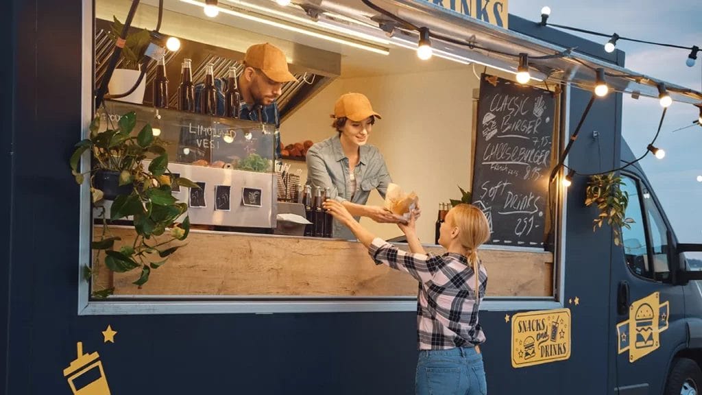 Two employees in yellow hats serve food to a customer out of a black food truck selling snacks and soft drinks.
