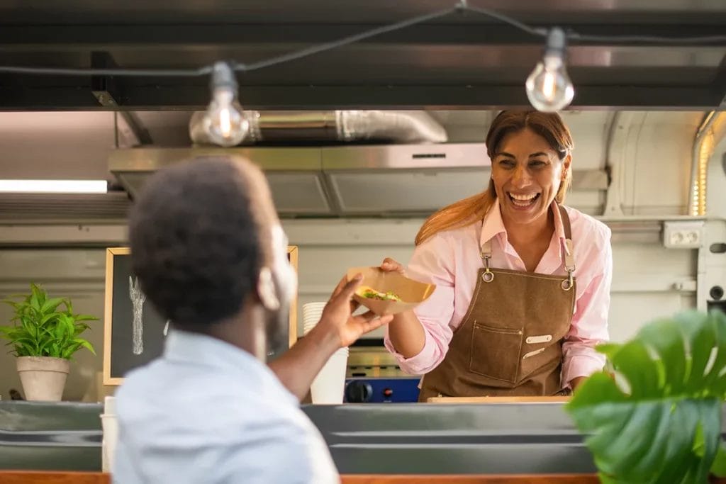 An employee in a food truck wearing a brown apron hands food to a customer from the service window.