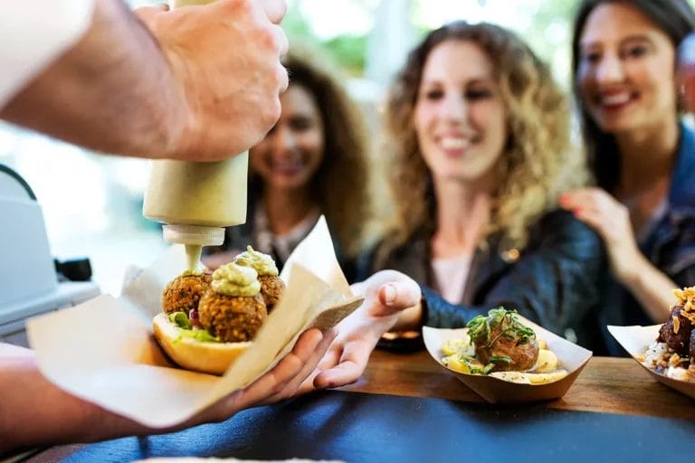 Three women wait for their orders at the service window of a food truck while the chef adds condiments to their dishes.