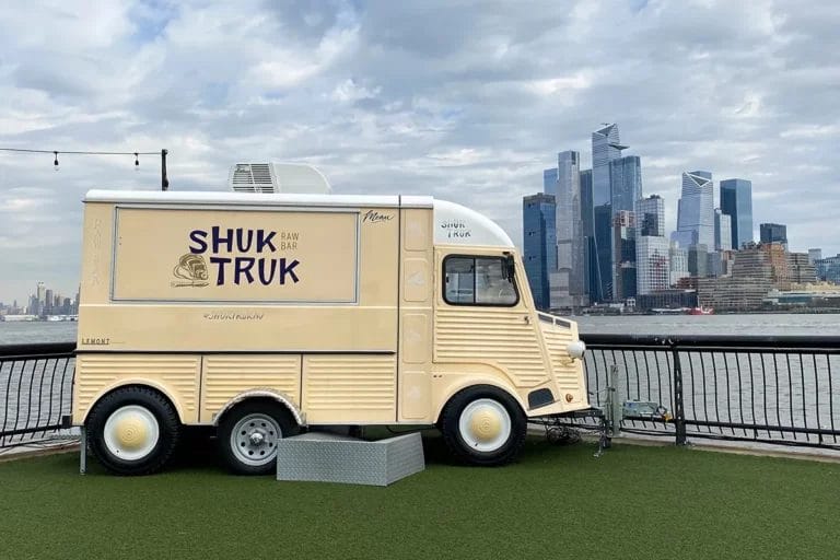 A food truck called the Shuk Truk parked along the Hudson River in Hoboken, New Jersey overlooking the Manhattan skyline.