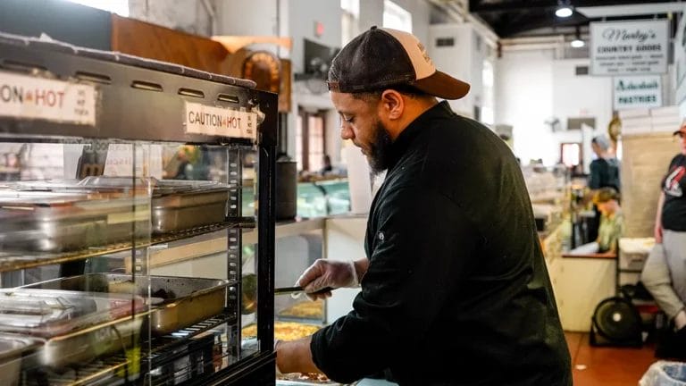 Corey Woods wears a black chef's coat and a backwards baseball cap while dishing up macaroni and cheese for a customer at his farmers market booth.