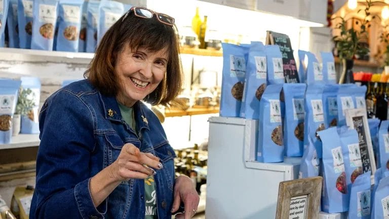 Mary Beth Shenk, salesperson at Happiness is... Granola, smiles at the camera while standing at the register.