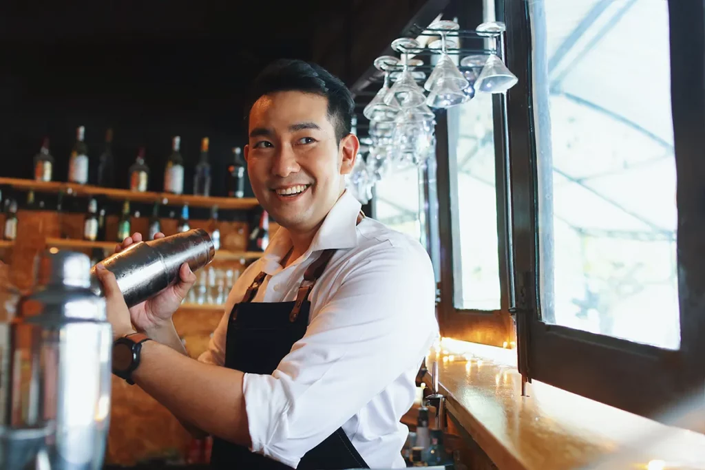 A male bartender in a white shirt and black apron smiles while holding a cocktail shaker at a bar.