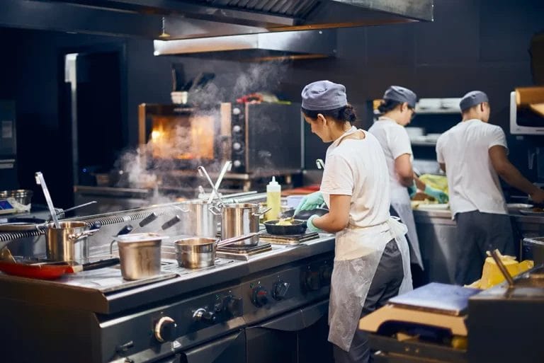Three chefs working together in a commercial kitchen, with one cooking food in a pan and the others working at a prep table.