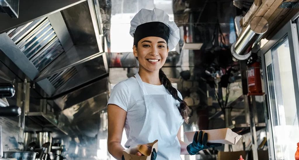 A woman in a chef's hat and apron smiles inside a food truck while holding two paper trays of food.