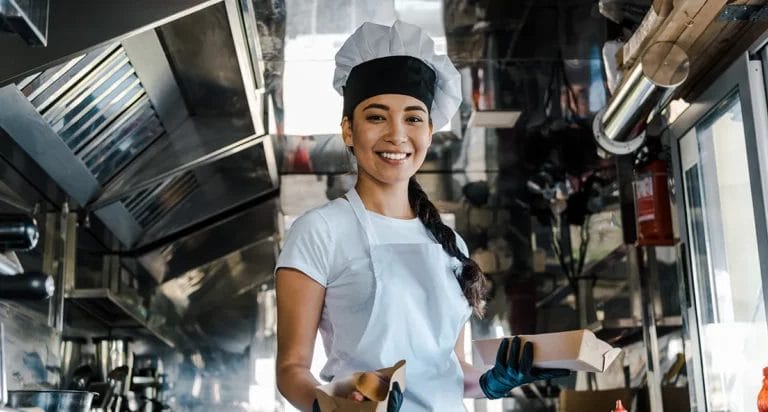 A woman in a chef's hat and apron smiles inside a food truck while holding two paper trays of food.