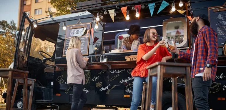 A group of people enjoying food on a sunny day in front of a black food truck.