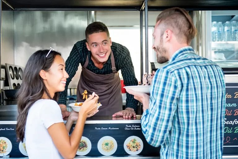 A worker smiles at two customers from inside a food truck after serving them food.