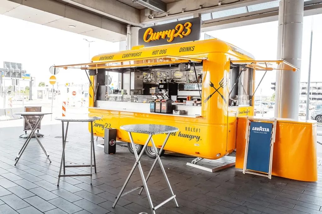 A yellow food trailer parked at a venue selling bratwurst, currywurst, hot dogs, and drinks.