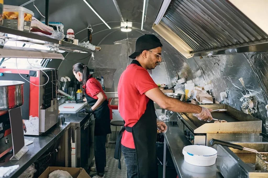 Two employees in red shirts and black aprons prepare food inside a food trailer.